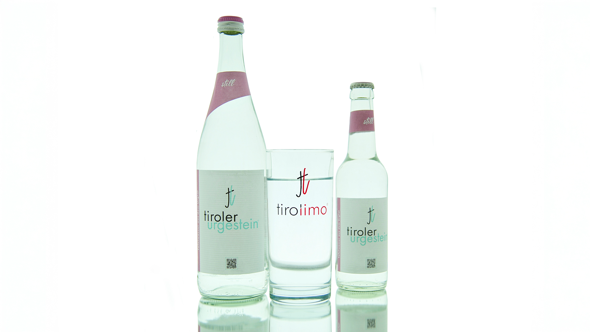 Zwei „tiroler urgestein still“-Wasserflaschen mit Trinkglas in der Mitte, auf spiegelndem Untergrund stilvoll arrangiert.