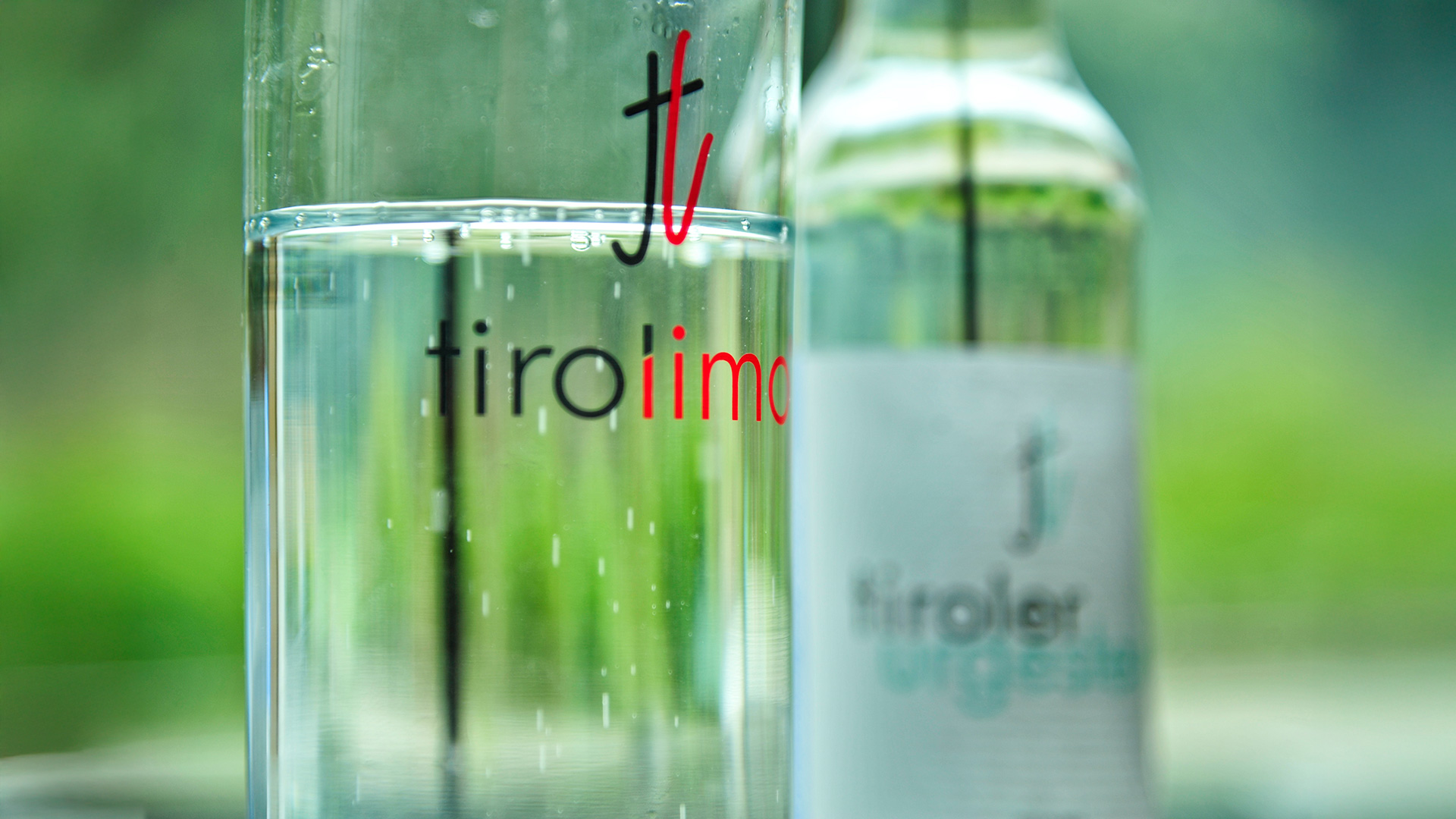 Nahaufnahme eines Glases mit „tiroliimo“-Schriftzug, gefüllt mit Wasser, daneben unscharfe „tiroler urgestein“-Flasche im Hintergrund.