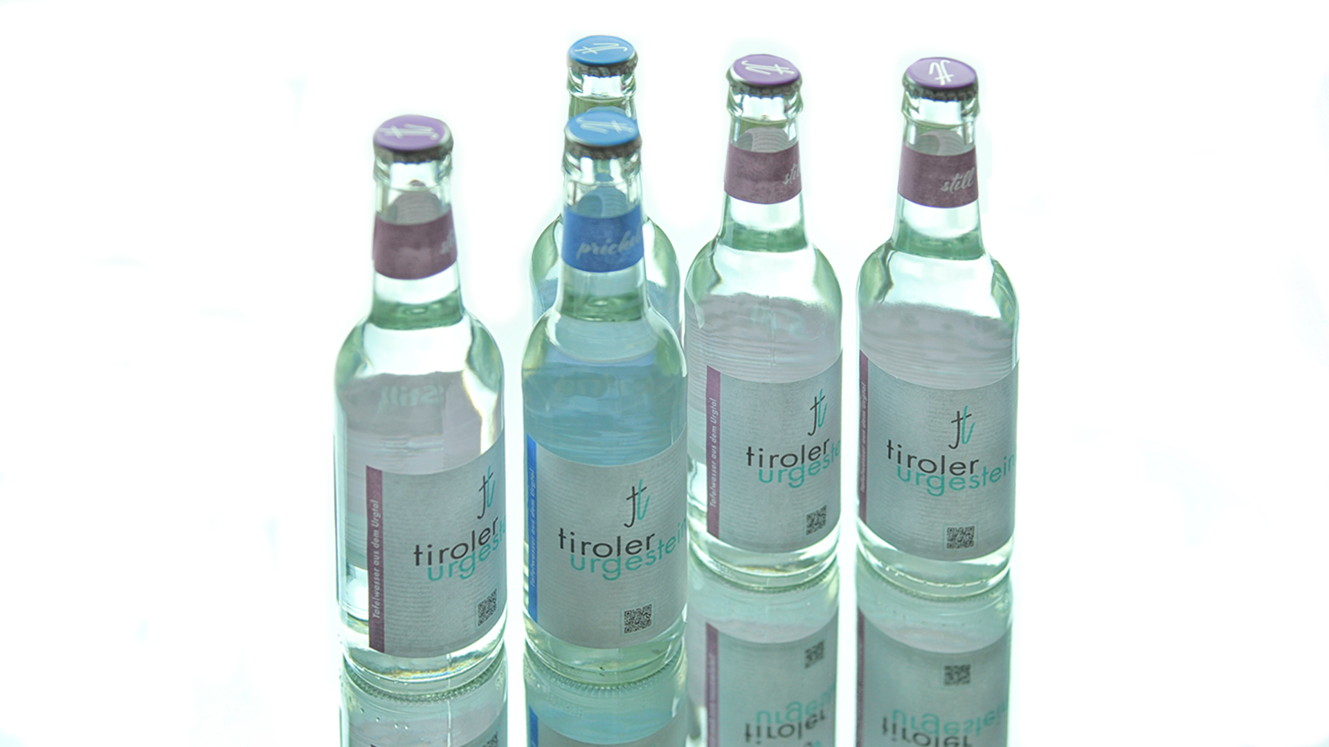 Fünf „tiroler urgestein“-Glasflaschen (still & prickelnd) auf spiegelndem Untergrund, aufrecht in Reihung dargestellt.